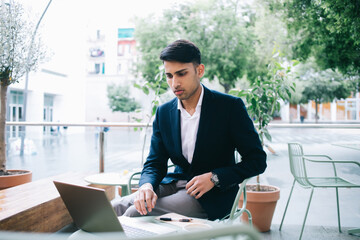 Serious hindu businessman in trendy formal wear watching video on laptop computer during distance job, pensive handsome prosperous male entrepreneur checking news from exchange browse on terrace