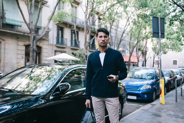 Young ethnic man with mobile phone and bag strolling on street in modern city