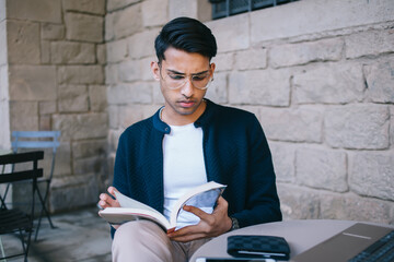 Concentrated young ethnic man reading book in front of laptop in cozy cafe