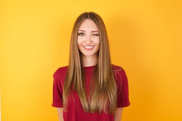 Fototapeta premium Close-up studio shot of positive coquettish Beautiful Young beautiful caucasian girl wearing red t-shirt , smiling happily, blinking at camera in a playful manner, flirting with you. 