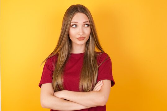 Photo of cheerful positive Beautiful Young beautiful caucasian girl wearing red t-shirt over isolated yellow background, looking aside into empty space thoughtful isolated over yellow background