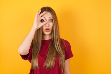Beautiful Young beautiful caucasian girl wearing red t-shirt over isolated yellow background doing ok gesture shocked with surprised face, eye looking through fingers. Unbelieving expression.