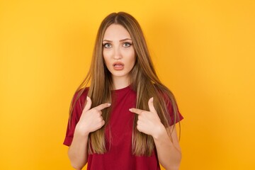 Photo of embarrassed Beautiful Young beautiful caucasian girl wearing red t-shirt over isolated yellow background indicates at herself with puzzled expression, being shocked 