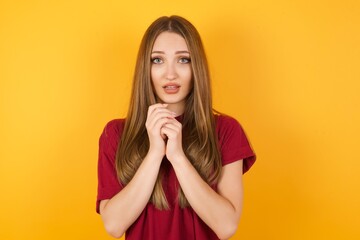 Beautiful Young beautiful caucasian girl wearing red t-shirt over isolated yellow wall smiles happily, glad to receive pleasant news from interlocutor, keeps hands together. People emotions concept.