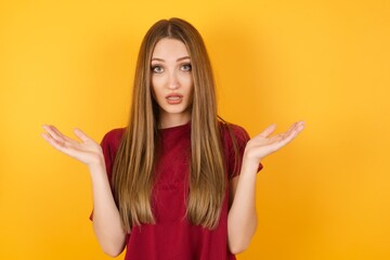 Surprised terrified Beautiful Young beautiful caucasian girl wearing red t-shirt over isolated yellow Gestures with uncertainty, stares at camera, puzzled as doesn't know answer on tricky question, 