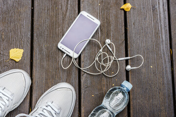 White sneakers, bottle of water, mobile phone and headphones, on the wooden floor outdoors.