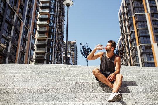 Young man exercising outside. Picture of athletist drinking water ad sitting on steps outside. Urban view. Body hydration. Big tall apartment buildings.