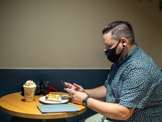 Shot of a man in a medical mask in a cafe  looking at his  phone - new normal