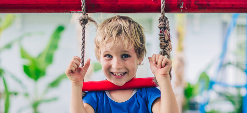 Close portrait Happy smile freckle blue eyed cute blond little boy sitting on a rope ladder, playing outside on playground in kindergarten. Daycare, activity, simple joys of childhood, summer