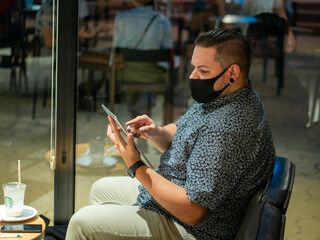 Shot of a man in a medical mask in a cafe  looking at his  tablet - new normal