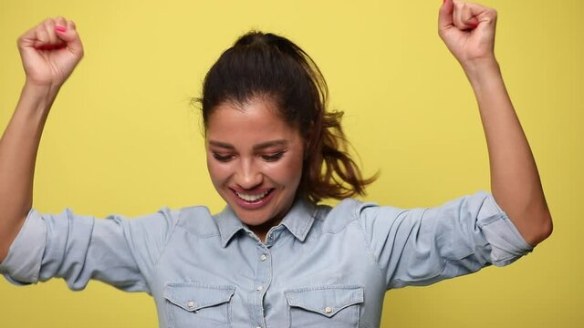 enthusiastic casual girl in blue jeans shirt, screaming, holding fists in the air and cheering, jumping and dancing on yellow background