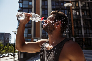 Young man exercising outside. Guy pouring some fresh water out of bottle on his face or splashing it. Stand alone in front urban apartment buildings. After exercising or training.