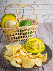 Guacamole with potato chips in plate and avocados in basket on wooden table