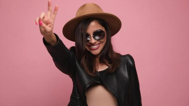 sexy woman in leather jacket wearing hat and sunglasses walking making peace gesture, dancing, arranging hair, making a pirouette, smiling and posing on pink background