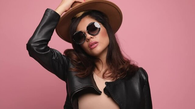 sexy cool woman wearing sunglasses and leather jacket, putting sensually hat on head, looking down and posing on pink background