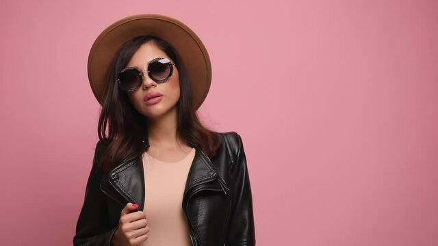 confident young woman in black leather jacket holding hand on hat and jacket, looking to side and posing, crossing arms, holding hand in a fashion pose, looking down on pink background