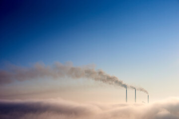Tops of three smoking stacks of thermal power station on the horizon taken from the hill, pipes in morning fog on blue sky, concept of energy generation, ecology and environmental pollution