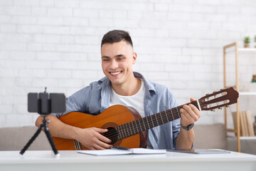 Guitar lesson and online blog. Young happy guy tutor plays on instrument and looks at webcam on smartphone and shoots video for students for study at home