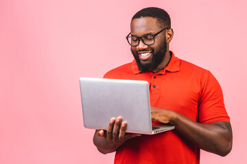 Young smiling african american man standing and using laptop computer isolated over pink background.