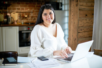 Beautiful young female accountant with curvy body and chubby cheeks typing on laptop, making...