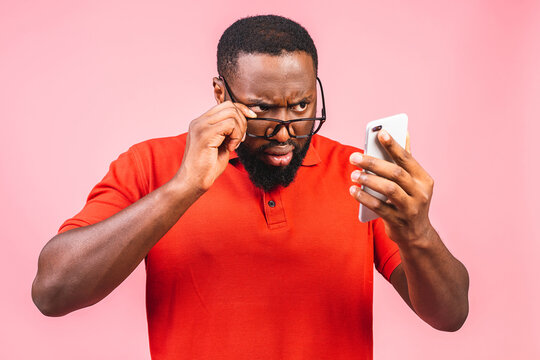 Closeup Portrait Of Handsome Young Man, Shocked, Surprised, Wide Open Mouth, Mad By What He Sees On His Cell Phone, Isolated On Pink Background. Negative Human Emotions, Facial Expressions, Feelings.