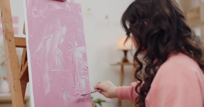 Backside view of young girl using modern painting technology without brushes and oil. Teenager painting with palette knife while sitting in front of molbert.Concept of creativity.