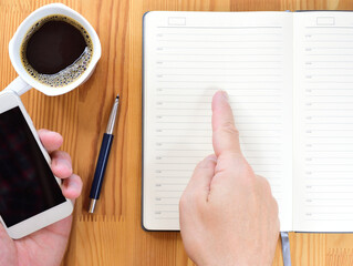 Diary opened on a blank page. Man's hands are visible. One hand is holding a smartphone, and the another is pointing to a diary page. Nearby is a mug of coffee.