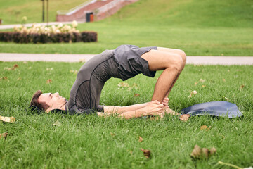 Young athletic man in sportswear doing yoga in the park. Practice asana outdoors. People exercising on green grass with yoga mat. Strong mature caucasian man in shoulder pose Kandharasana