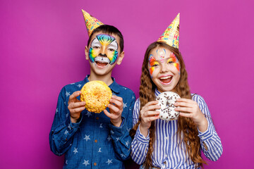 Two happy little best friends with aquagrim wear birthday cap holding donut isolated on violet background. Birthday concept