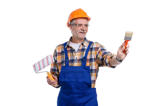 Portrait of positive senior male builder holding paint roller  and brush in hand, wearing special uniform, apron and protective helmet, isolated on white background