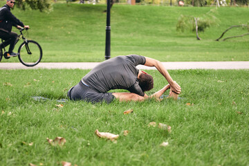 Young athletic man in sportswear doing yoga in the park. Practice asana outdoors. People exercising on green grass with yoga mat. Man in Parivritti Janu Sirshasana spiralled head to knee pose