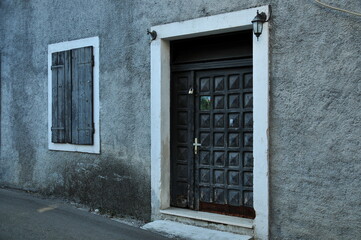 A black wooden door with pattern of the cut out squares, framed by the white doorway and threshold, against a gray textured wall with closed dark gray shutters.