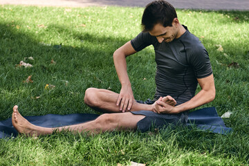 Young athletic man in sportswear doing yoga in the park. Practice asana outdoors. People exercising stretching on green grass with yoga mat. Fitness and healthy lifestyle. Concept of meditation