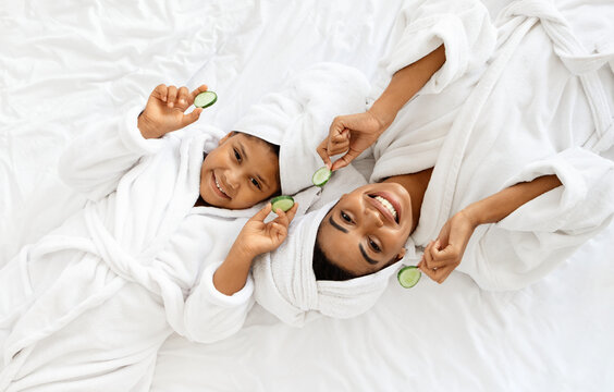 Home Spa. Happy African Mom And Daughter In Bathrobes With Cucumber Slices