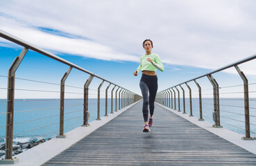 Strong female jogger in stylish tracksuit enjoying morning run across seashore boardwalk, Caucasian...