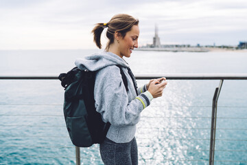 Focused adult woman with backpack using smartphone on waterfront