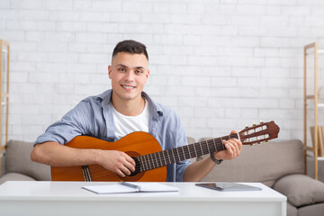 Obraz premium Online music lessons. Cheerful guy with guitar sits on couch in living room interior and records video or makes record for modern blog