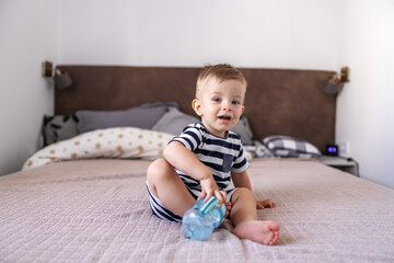 Cute little blond boy smiling and looking at camera with his beautiful blue eyes while sitting on bed in bedroom and holding his bottle with water.