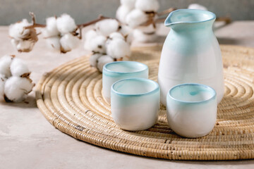 Fototapeta na wymiar Sake ceramic set for traditional japanese alcohol drink rice wine sake, pitcher and three cups, standing on straw napkin with cotton flowers over beige texture background.