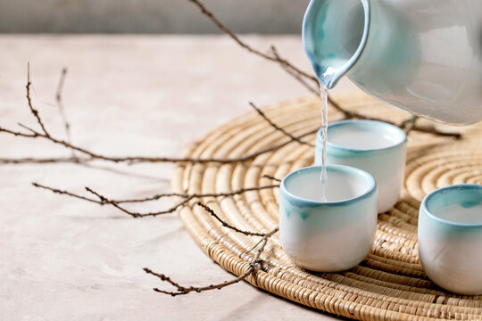 Sake ceramic set for traditional japanese alcohol drink rice wine sake pouring from pitcher in three cups, standing on straw napkin with dry branches over beige texture background.