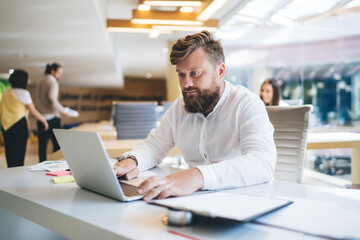 Caucasian bearded male entrepreneur working remotely with program code for system updation, smart casual businessman messaging with corporate partners while making web banking on laptop computer
