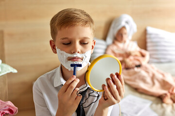 adorable kids couple have happy morning, take care of themselves, boy is shaving beard with shaving foam on face, at home on bed, girl with towel on head saws the nail next to him