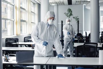 People in hazmat, protective mask and glasses making disinfection of furniture in office with chemicals during coronavirus quarantine - obrazy, fototapety, plakaty