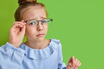 close-up portrait of little caucasian girl in eyeglasses, she is attentively looking at camera, in blue blouse, isolated on green background