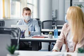 Obraz premium Social distance of workers in office during quarantine. Guy and woman in protective masks are talking, sitting at tables with computers and antiseptics