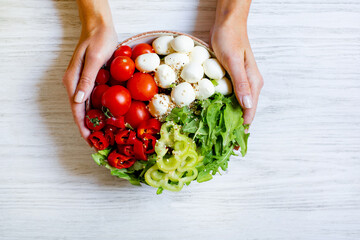 plate with vegetables tomatoes pepper arugula cheese mazzarella in female hands on a white wooden table top view