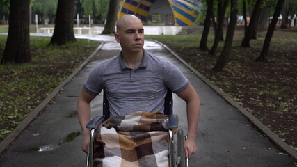 A young man with oncology rides a wheelchair through the park. The man is bald due to chemotherapy.
