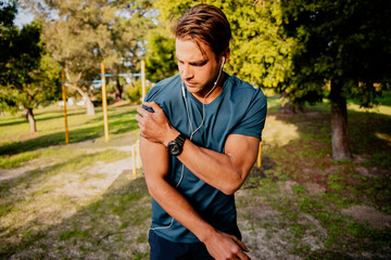 Obraz premium Injured young male athlete looking concerned holding shoulder in pain after exercising outdoors with smartwatch and ear phones