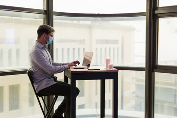 man teleworking from home with his laptop and wearing a mask, in front of a large window.