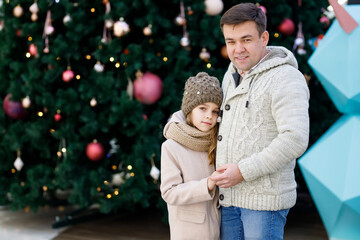 dad with a little girl in a coat, hat and scarf near a large Christmas tree in a shopping center. family walks during the new year holidays. happy childhood.